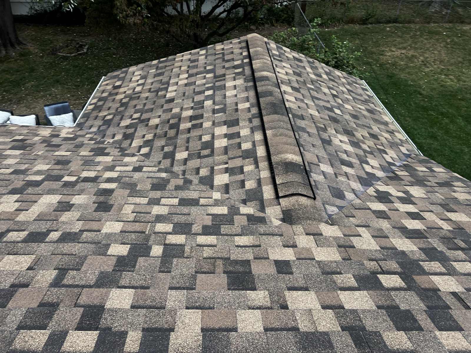 Looking down at the hip of a newly laid brown, tan and charcoal multi tone architectural shingle roof, capped with ridge cap shingles running up the hip and carrying a dark ridge vent strip along the ridge, with the two roof planes falling away either side and a mown lawn, chain link fence and cushioned outdoor furniture below