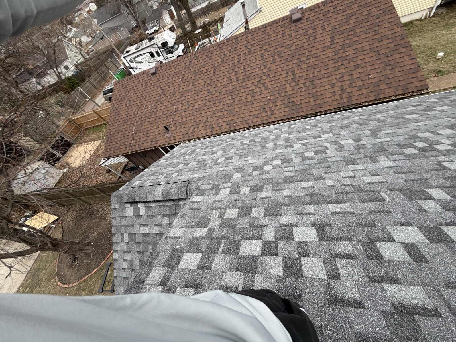 A newly laid grey and charcoal architectural shingle roof in the foreground with a ridge vent along its hip, meeting the old brown shingle roof of the adjoining garage, above a winter back yard with a paver path, a wooden fence, a white recreational vehicle and a skip in the alley