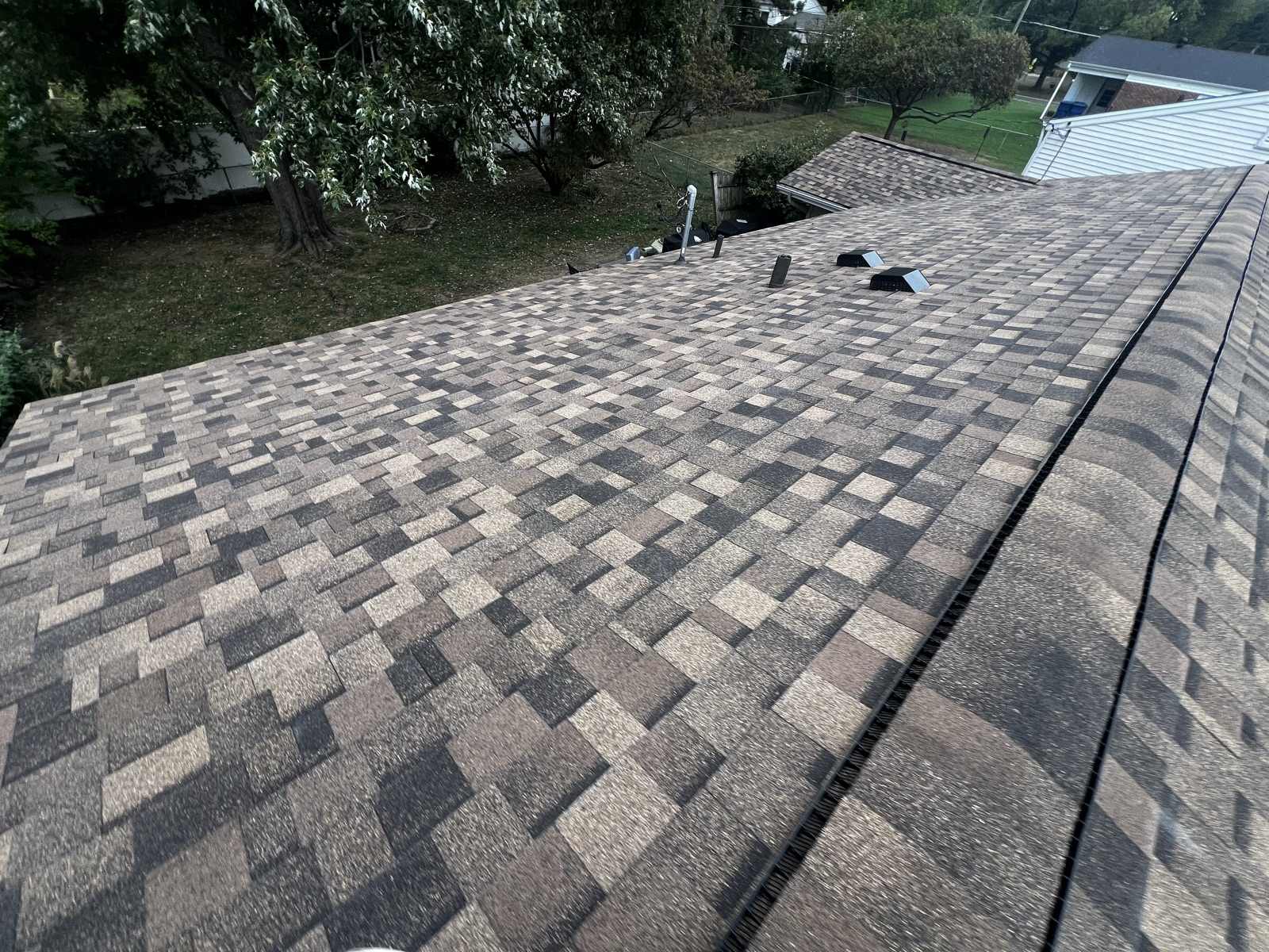 A wide plane of newly laid brown, tan and charcoal multi tone architectural shingles with the capped ridge and a dark ridge vent running down the right side, two black box vents and a small metal flue near the far end of the ridge, and a back yard with a large tree and chain link fence below