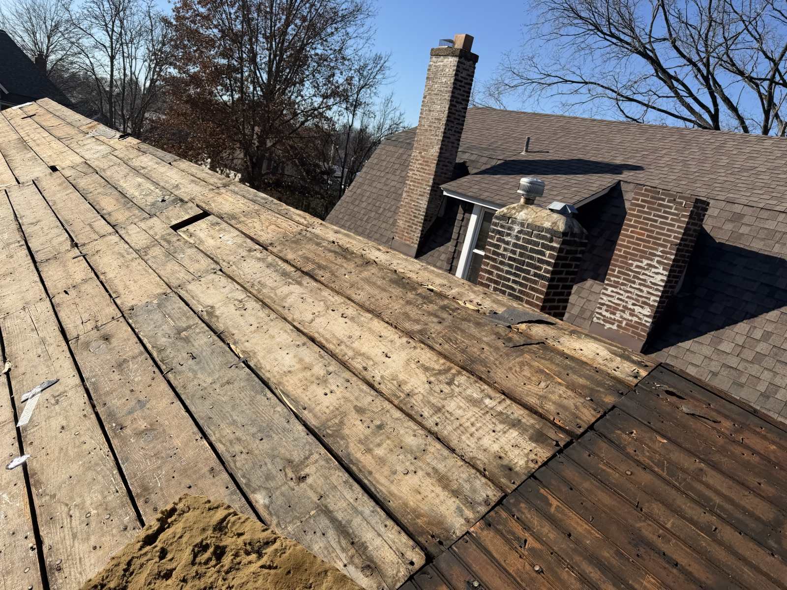 A roof plane stripped back to bare wide plank decking studded with old nails running up the left of the frame, with the main house behind still carrying its weathered brown shingles, two brick chimneys with visible spalling and missing mortar, a small dormer with a metal turbine vent, bare winter trees and a clear blue sky