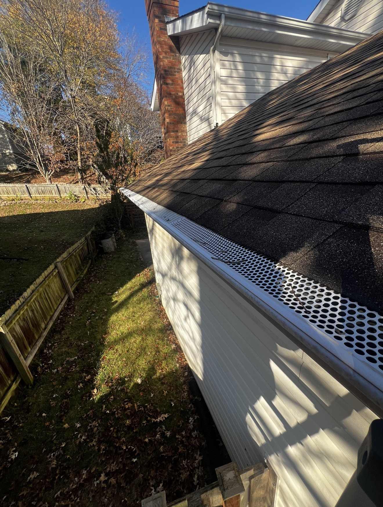 Looking along the eave of a weathered dark brown shingle roof with a white perforated metal gutter guard punched with round holes fitted the length of the gutter, two bare twigs and a few small dark scraps of debris lying on top of the perforations, and white vinyl siding, a red brick chimney, a wooden privacy fence, a mown lawn and fallen autumn leaves below under a clear blue sky