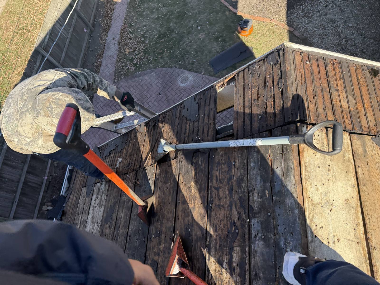 Stripped dark plank decking seen from above with an orange handled tear off bar, an aluminium D handle roofing shovel and two red flat spades lying across it, an aluminium ladder hooked over the eave, and a worker in a camouflage jacket crouched at the roof edge at the upper left